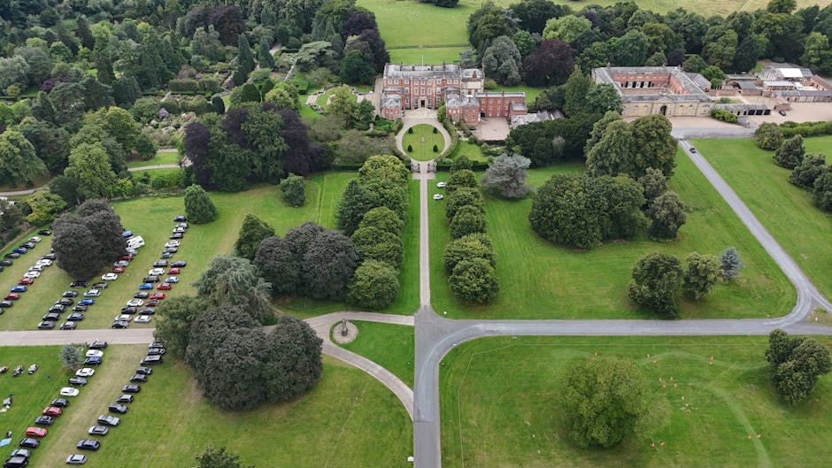 An aerial view of a landscaped estate with a large, historic brick and stone mansion at the top center, surrounded by well-maintained lawns, gravel pathways, and a variety of mature trees, including s