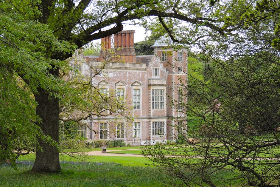 A historic brick mansion with multiple large windows and ornate architectural details, partially obscured by mature trees with fresh green foliage. The foreground features a well-maintained garden wit